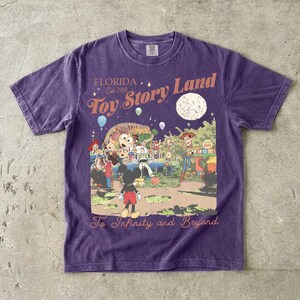 May include: A purple short-sleeve t-shirt featuring a graphic print with the text Florida Est 2018 Toy Story Land and To Infinity and Beyond. The illustration depicts Mickey Mouse, Woody, Buzz Lightyear, and other characters in a park setting.