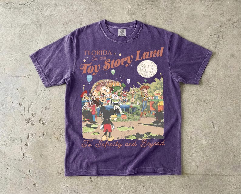 May include: A purple short-sleeve t-shirt featuring a graphic print with the text Florida Est 2018 Toy Story Land and To Infinity and Beyond. The illustration depicts Mickey Mouse, Woody, Buzz Lightyear, and other characters in a park setting.