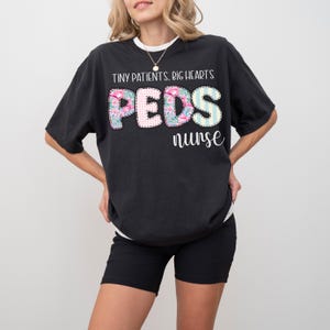 Op de afbeelding: Zwart T-shirt met de tekst "TINY PATIENTS. BIG HEARTS" boven het woord "PEDS" in een bloemen-, ruit- en streepontwerp, en het woord "nurse" eronder. Het shirt wordt gedragen met een zwarte short.