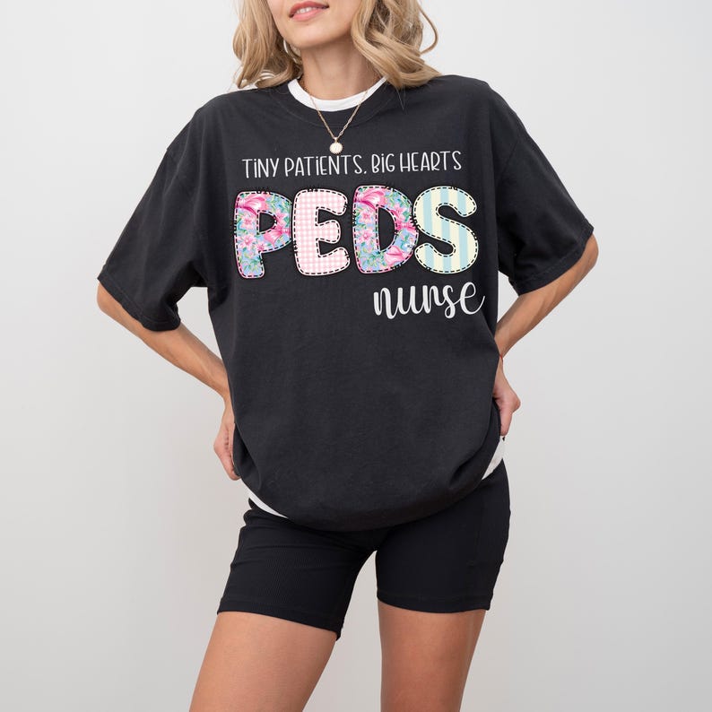 Op de afbeelding: Zwart T-shirt met de tekst "TINY PATIENTS. BIG HEARTS" boven het woord "PEDS" in een bloemen-, ruit- en streepontwerp, en het woord "nurse" eronder. Het shirt wordt gedragen met een zwarte short.