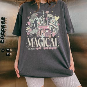 May include: Dark gray t-shirt with a colorful graphic design featuring a castle, Mickey Mouse ears, and the text "The Most Magical Place on Earth." The design includes pink, yellow, and blue elements. The shirt is worn by a person.