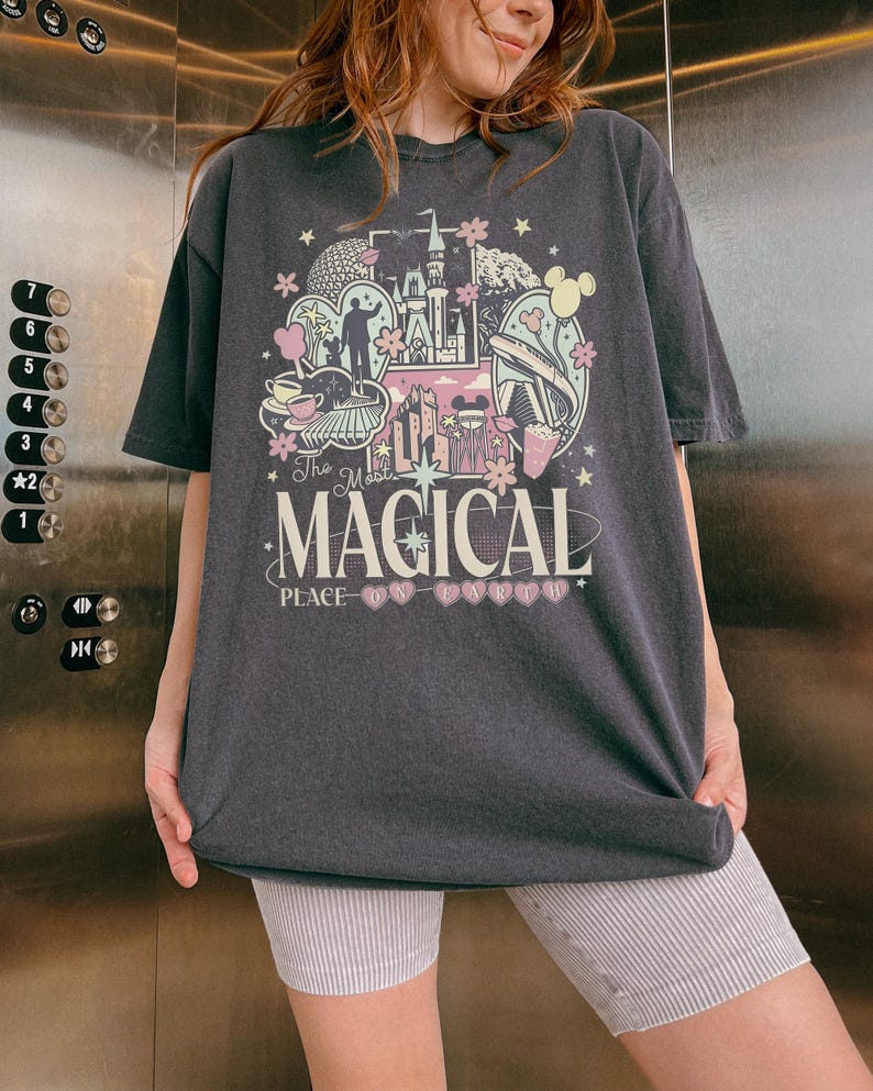 May include: Dark gray t-shirt with a colorful graphic design featuring a castle, Mickey Mouse ears, and the text "The Most Magical Place on Earth." The design includes pink, yellow, and blue elements. The shirt is worn by a person.