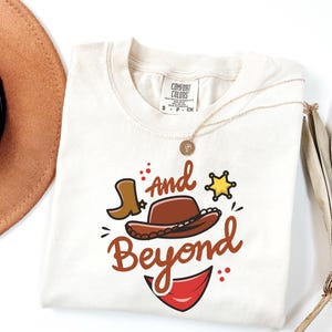 May include: Cream-colored t-shirt with a cowboy-themed graphic. The design includes a brown cowboy hat, a boot, a sheriff's star, and the words "and Beyond" in a playful font. A gold necklace is draped on the shirt.
