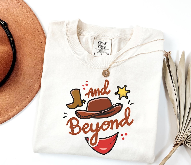 May include: Cream-colored t-shirt with a cowboy-themed graphic. The design includes a brown cowboy hat, a boot, a sheriff's star, and the words "and Beyond" in a playful font. A gold necklace is draped on the shirt.