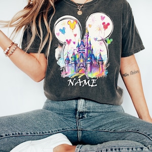 May include: Dark gray t-shirt featuring a colorful castle design within Mickey Mouse ear outlines. The design includes balloons and fireworks. The word "NAME" is printed below the castle. The shirt is paired with blue jeans and white shoes.