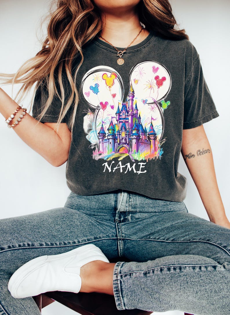 May include: Dark gray t-shirt featuring a colorful castle design within Mickey Mouse ear outlines. The design includes balloons and fireworks. The word "NAME" is printed below the castle. The shirt is paired with blue jeans and white shoes.