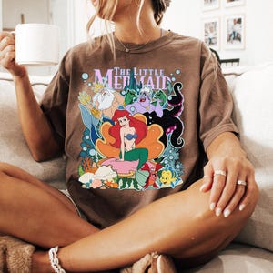 May include: Brown t-shirt featuring a colorful graphic of Ariel and other characters from "The Little Mermaid". The shirt has the text "The Little Mermaid" above the graphic. The person is sitting on a beige couch.