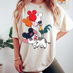 May include: Cream-colored t-shirt featuring a cartoon illustration of a pig wearing a Mickey Mouse hat, a rooster, and a cow. The pig is riding the cow, and the rooster is standing on the cow's back. Balloons in red, orange, pink, and white are floating above.