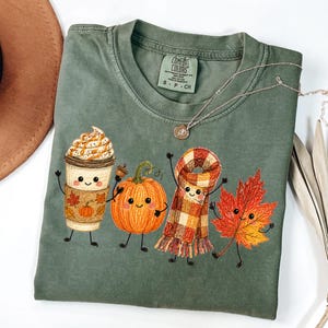 May include: An olive green t-shirt featuring a graphic of a smiling coffee cup, a pumpkin, a plaid scarf, and a maple leaf. The shirt has a Comfort Colors label at the neck. The graphic includes a coffee cup with whipped cream, a round orange pumpkin, a brown and orange plaid scarf, and a vibrant red and orange maple leaf, all with small black arms and legs.