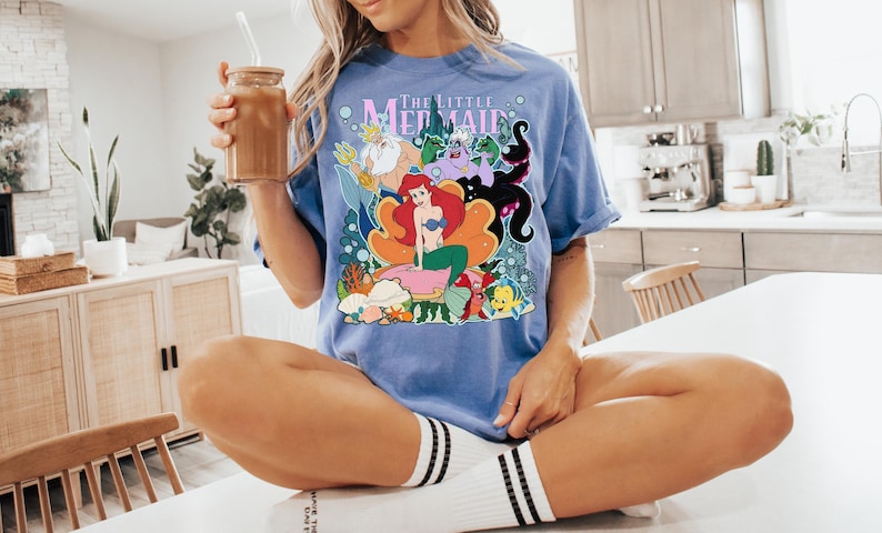 May include: A light blue t-shirt featuring a colorful graphic from "The Little Mermaid." The design includes Ariel, King Triton, Ursula, and other characters. The shirt is worn by a person sitting on a white surface.