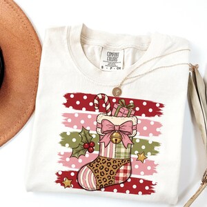 May include: A cream-colored t-shirt with a Comfort Colors label featuring a graphic of a Christmas stocking. The stocking has a leopard print, plaid, and striped pattern, adorned with a pink bow, a bell, a candy cane, and a gift box.