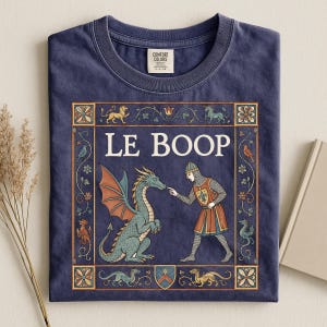 May include: A navy blue t-shirt featuring a medieval-style graphic with the text LE BOOP. The design shows a knight in chainmail touching the nose of a green dragon, surrounded by a decorative border with floral motifs and small mythical creatures.
