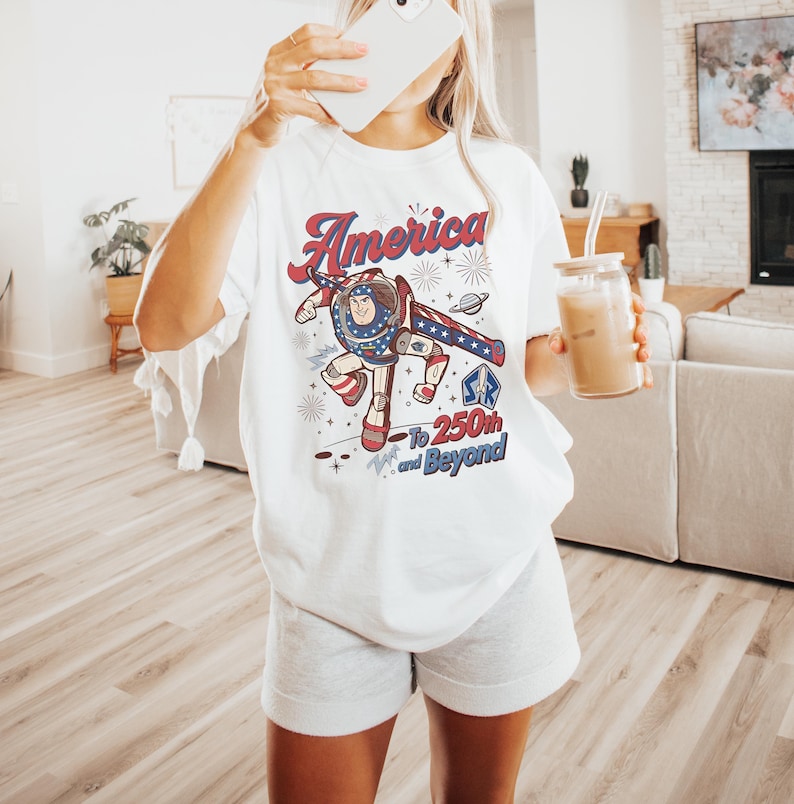May include: A white t-shirt featuring a graphic of a space ranger character in a red, white, and blue suit. The text reads America To 250th and Beyond, accompanied by star and firework illustrations. The design includes a small SR logo emblem.
