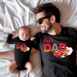 May include: Black t-shirts and a baby bodysuit with a race car theme. The adult's shirt says "Fast Dad" with a checkered flag and a red race car. The baby's bodysuit says "Fast Like Dad" with a similar design.