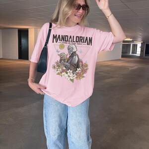 以下が含まれることがあります： 淡いピンク色の半袖Tシャツ。マンダロリアンとグローグーが色とりどりの花や蝶に囲まれたグラフィックプリントが施されています。中央のイラストの上部には、黒い文字で「MANDALORIAN THIS IS THE WAY」と記されています。