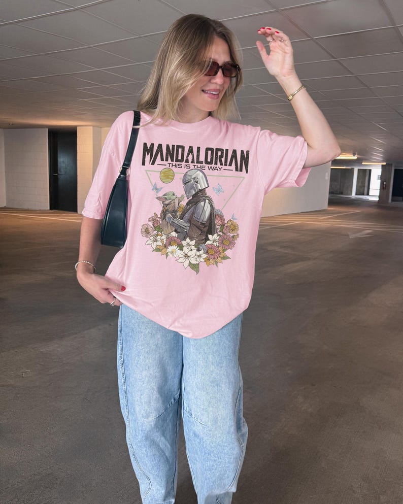 以下が含まれることがあります： 淡いピンク色の半袖Tシャツ。マンダロリアンとグローグーが色とりどりの花や蝶に囲まれたグラフィックプリントが施されています。中央のイラストの上部には、黒い文字で「MANDALORIAN THIS IS THE WAY」と記されています。