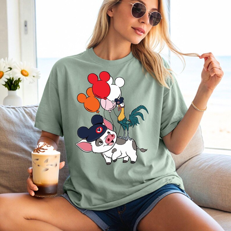 May include: Sage green t-shirt featuring a cartoon pig wearing a hat and holding balloons shaped like Mickey Mouse. A rooster stands on the pig's back. The shirt is a relaxed fit.