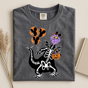 Puede incluir: Una camiseta gris carbón con un gráfico de un dragón morado con un disfraz de esqueleto negro. El dragón sostiene tres globos naranjas con siluetas de orejas de ratón negras y un pequeño cubo de calabaza naranja lleno de dulces.