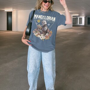 以下が含まれることがあります： スレートブルーの半袖Tシャツ。マンダロリアンとグローグーが花や蝶に囲まれたグラフィックプリントが施されています。テキストには「MANDALORIAN THIS IS THE WAY」と書かれています。ライトブルーのデニムジーンズと白いスニーカーを合わせています。