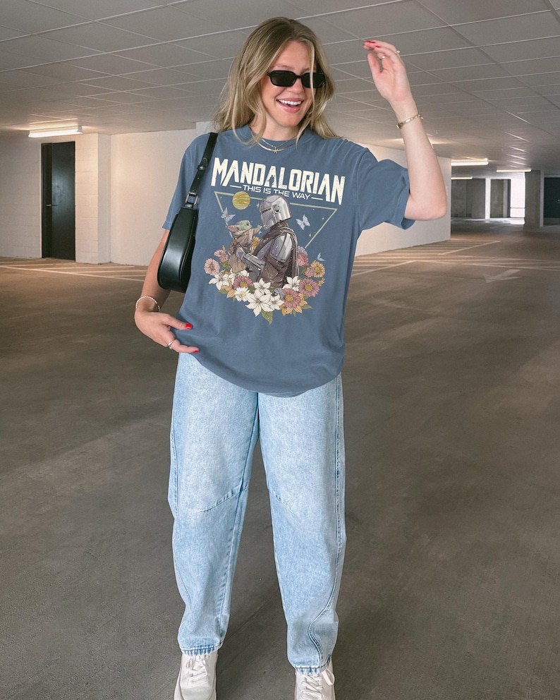 以下が含まれることがあります： スレートブルーの半袖Tシャツ。マンダロリアンとグローグーが花や蝶に囲まれたグラフィックプリントが施されています。テキストには「MANDALORIAN THIS IS THE WAY」と書かれています。ライトブルーのデニムジーンズと白いスニーカーを合わせています。