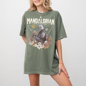 以下が含まれることがあります： オリーブグリーンのTシャツ。マンダロリアンとグローグー、色とりどりの花、蝶、黄色い太陽のグラフィックプリントが施されています。キャラクターの上部には、かすれた白のフォントで「MANDALORIAN」と「THIS IS THE WAY」の文字がプリントされています。