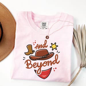 May include: A light pink t-shirt with a graphic design featuring a cowboy hat, boot, sheriff's star, and the words "And Beyond" in brown script. A gold necklace with a pendant is draped on the shirt.