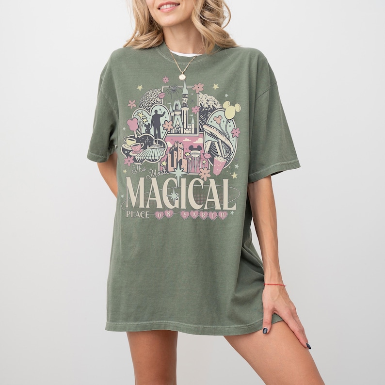 May include: Olive green t-shirt featuring a colorful graphic design with the text "The Most Magical Place". The design includes a castle, flowers, and other whimsical elements. The shirt has short sleeves and a relaxed fit.