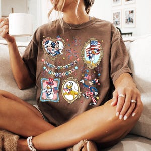 May include: Brown t-shirt featuring cartoon characters in patriotic frames. The shirt has the text "250th Anniversary 1776-2026" and is worn by a person sitting on a couch.