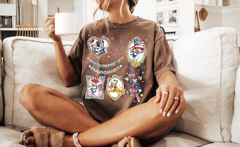 May include: Brown t-shirt featuring cartoon characters in patriotic frames. The shirt has the text "250th Anniversary 1776-2026" and is worn by a person sitting on a couch.