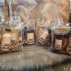 May include: Several clear plastic tote bags filled with apothecary products. Each bag contains a variety of items, including bottles, jars, and small packages, all with labels featuring the text "Mystical Mrs. T's Apothecary". The bags are arranged on a reflective surface.