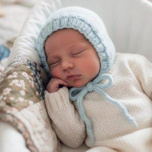 Può includere: Un bambino che dorme con un cappuccio di maglia azzurra con un fiocco abbinato e un maglione color crema. Il bambino è adagiato in una culla bianca, appoggiato su una coperta a fantasia floreale.