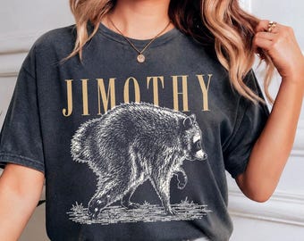May include: A charcoal gray short-sleeve t-shirt featuring the text JIMOTHY in a tan serif font above a detailed black and white graphic illustration of a raccoon walking on a textured ground surface, with a crew neckline and a relaxed fit.