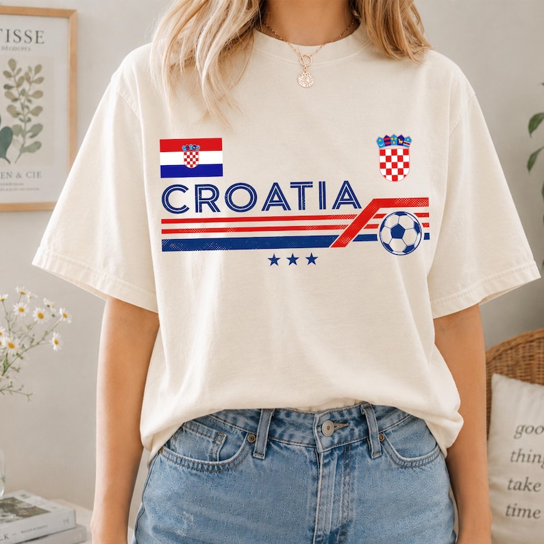 May include: A cream-colored short-sleeve t-shirt featuring the text CROATIA in blue, a red and white checkered flag, the Croatian coat of arms, a soccer ball graphic, and three blue stars above horizontal red and blue stripes on the front.