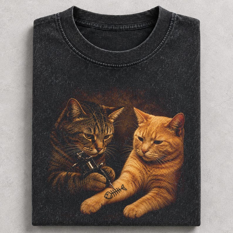 May include: Dark gray t-shirt featuring a humorous illustration of two cats. One cat is tattooing a fishbone on the other cat's arm. The design is printed on a vintage-style, crew neck tee.