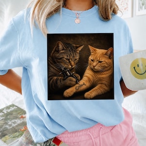 May include: Light blue sweatshirt featuring a graphic of two cats, one tattooing a fishbone on the other's arm. The image is framed in a square. A white mug with a yellow smiley face is visible.