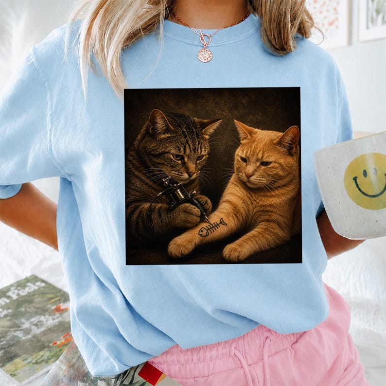 May include: Light blue sweatshirt featuring a graphic of two cats, one tattooing a fishbone on the other's arm. The image is framed in a square. A white mug with a yellow smiley face is visible.