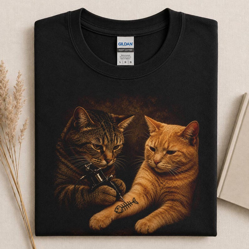 May include: Black Gildan heavy cotton sweatshirt featuring a humorous graphic of two cats. One cat is tattooing a fishbone on the other cat's arm. The sweatshirt has a crew neck and long sleeves. The shirt is a size large.