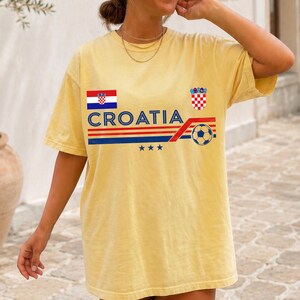 May include: A pale yellow short-sleeve t-shirt featuring the text CROATIA in blue, a horizontal red and blue striped graphic, a soccer ball icon, the Croatian flag, the Croatian coat of arms, and three small blue stars centered on the chest.