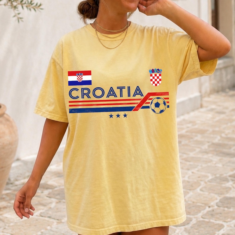 May include: A pale yellow short-sleeve t-shirt featuring the text CROATIA in blue, a horizontal red and blue striped graphic, a soccer ball icon, the Croatian flag, the Croatian coat of arms, and three small blue stars centered on the chest.