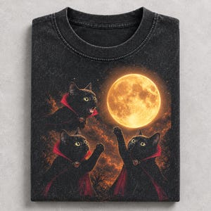 May include: A black acid-wash t-shirt featuring a graphic of three black cats wearing red-lined capes. The cats are positioned against a dark, cloudy sky with a large, glowing yellow full moon. One cat has its mouth open, while the others reach up.