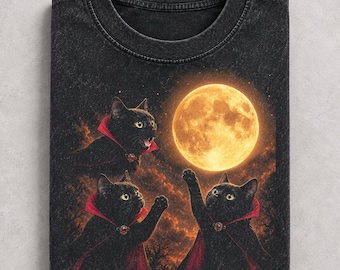 May include: A black acid-wash t-shirt featuring a graphic of three black cats wearing red-lined capes. The cats are positioned against a dark, cloudy sky with a large, glowing yellow full moon. One cat has its mouth open, while the others reach up.