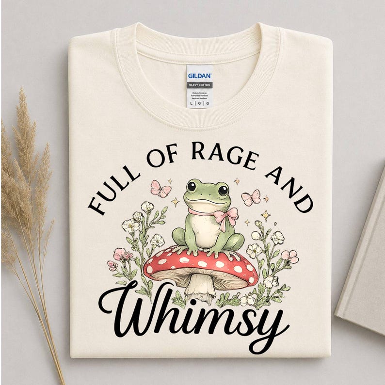 Puede incluir: Camiseta color crema con un dise&ntilde;o caprichoso. El gr&aacute;fico presenta una rana verde con un lazo rosa, sentada en una seta roja con lunares blancos, rodeada de flores y mariposas. El texto dice "FULL OF RAGE AND Whimsy."