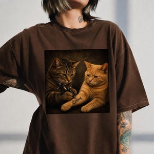 May include: Brown t-shirt featuring a square image of two cats. One cat is tattooing a fishbone on the other cat's arm. The cats are brown and orange. The t-shirt is a dark brown color.