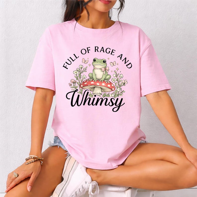 Puede incluir: Camiseta rosa claro con un dise&ntilde;o caprichoso. Presenta una rana verde sentada en una seta roja y blanca, rodeada de flores y mariposas. El texto "FULL OF RAGE AND Whimsy" est&aacute; impreso encima y debajo de la ilustraci&oacute;n.