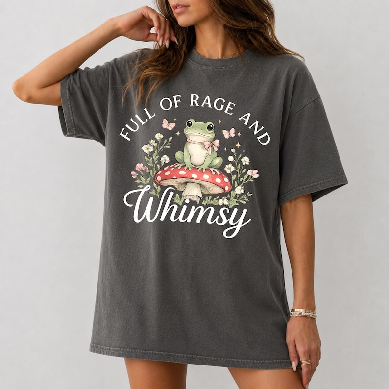 Puede incluir: Camiseta gris oscuro con un dise&ntilde;o caprichoso. El gr&aacute;fico incluye una rana verde con pajarita, sentada sobre una seta roja y blanca, rodeada de flores y mariposas. El texto dice "FULL OF RAGE AND Whimsy".