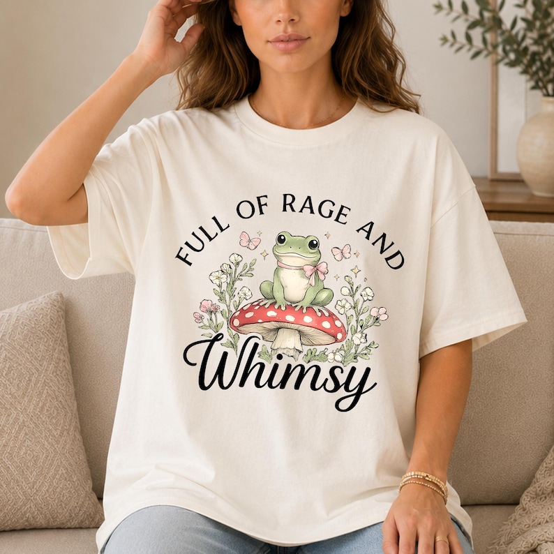 Puede incluir: Camiseta color crema con un gr&aacute;fico caprichoso. El dise&ntilde;o presenta una rana verde con un lazo rosa, sentada en una seta roja y blanca, rodeada de flores y mariposas. El texto dice "FULL OF RAGE AND Whimsy."