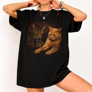 May include: Black t-shirt featuring a graphic of two cats. One cat is tattooing a fishbone on the other cat's arm. The cats are brown and orange, set against a dark background. The shirt has a relaxed fit.