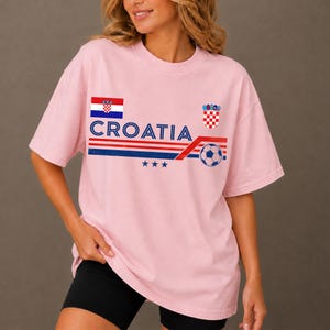 May include: A light pink short-sleeve t-shirt featuring the text CROATIA in blue, a red, white, and blue striped graphic, a soccer ball, three blue stars, the Croatian flag, and the Croatian coat of arms printed on the front chest area.