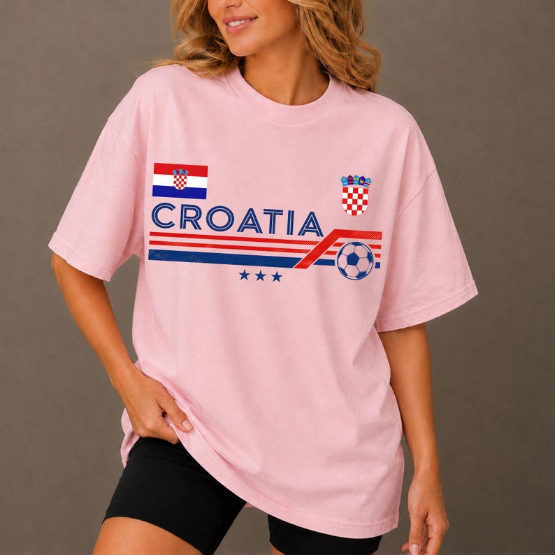 May include: A light pink short-sleeve t-shirt featuring the text CROATIA in blue, a red, white, and blue striped graphic, a soccer ball, three blue stars, the Croatian flag, and the Croatian coat of arms printed on the front chest area.
