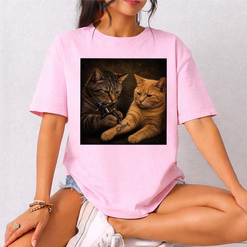 May include: A light pink t-shirt featuring a square graphic of two cats. One cat is tattooing the other cat's arm with a fishbone design. The cat doing the tattoo is brown and the cat getting the tattoo is orange.